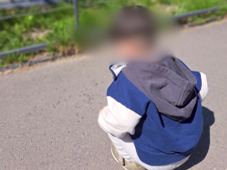 青いパーカーを着て、公園の地面にある何かをじーっと観察するためにしゃがみ込む男の子の後ろ姿
