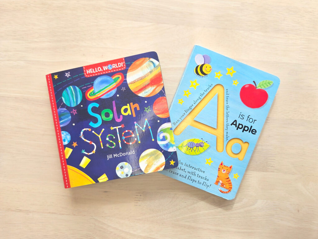 明るい木目調のテーブルの上に、英語の知育絵本「Hello World! Solar System」と「Hello World! ABCs」の2冊が並べて置かれている。左側は宇宙の惑星が描かれた紺色の表紙、右側は大きなアルファベットのAが描かれた水色の表紙で、どちらも色彩豊かで知育に最適なデザインであることが一目でわかる一枚。