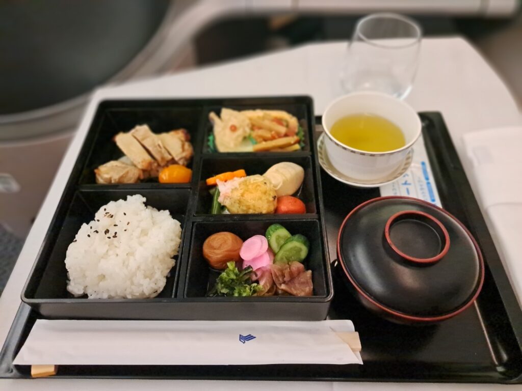 シンガポール航空ビジネスクラスで提供された、本格的な和風の機内食（花恋暦）。黒い松花堂弁当形式の器に、ご飯、焼き魚、煮物、お浸し、漬物などが美しく盛り付けられている。横にはお吸い物の椀、湯呑みに入った緑茶、グラスに入った水、箸袋が置かれている。