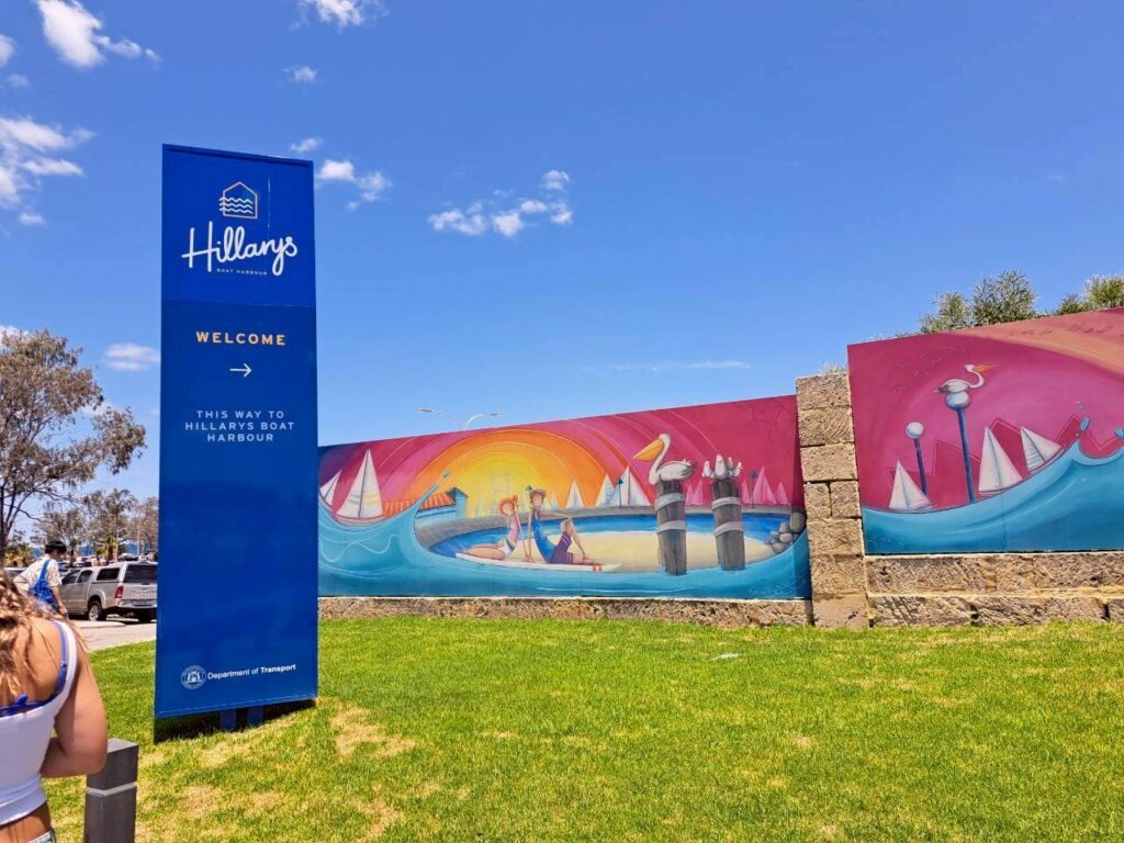 オーストラリア・パース北部の人気スポット「Hillarys Boat Harbour（ヒラリーズ・ボート・ハーバー）」の入り口。青い空の下、鮮やかな青色のウェルカム看板（「Hillarys」のロゴと「WELCOME」の文字）が立っている。その奥には、ヨットや海辺の様子が描かれたカラフルな壁画、緑の芝生、そして駐車場の一部が見える。
