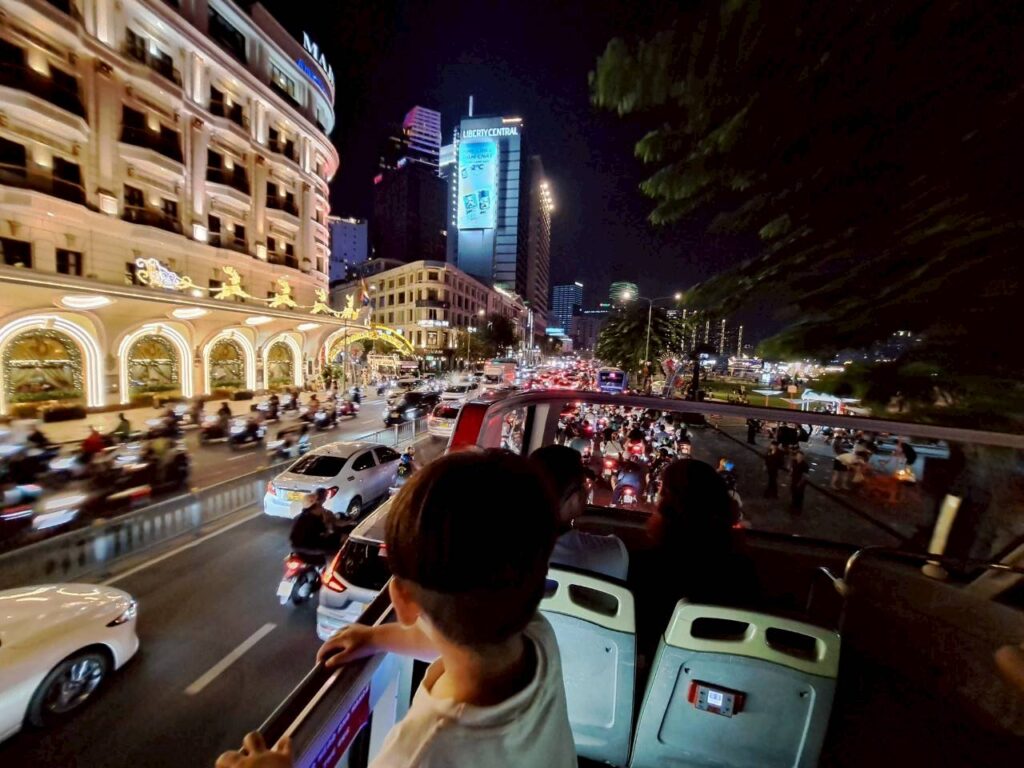 ベトナム　ホーチミン　異国の華やかな夜景の中を走る、オープントップの二階建てバスからの眺め。身を乗り出して光り輝く街並みを見つめる2歳の子供。