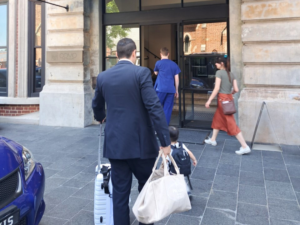 オーストラリア・パースの高級ホテル「COMO the Treasury（コモ・ザ・トレジャリー）」の入り口。石造りの重厚な建物の前で、紺色のスーツを着てスーツケースを引く男性と、白いキャンバストートバッグを持った小さな子供が背中を向けて立っている。隣にはホテルの真鍮製看板と青い車の一部が見える。