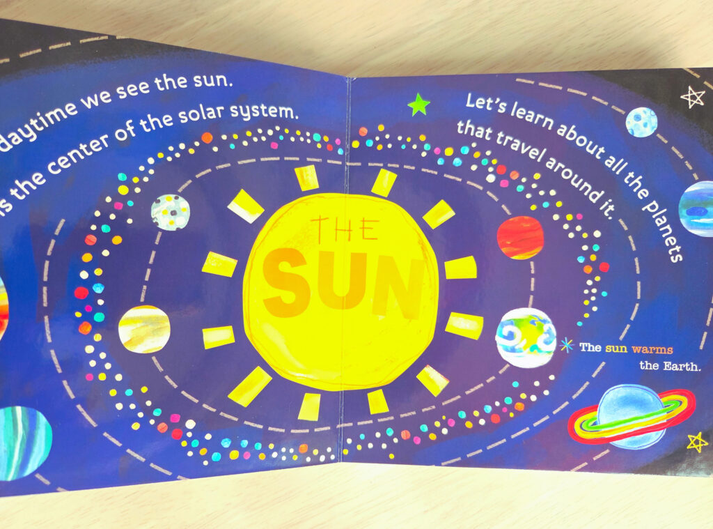 色鮮やかな太陽系の惑星が描かれたボードブック「Hello World! Solar System」の見開き。中央には「THE SUN」と書かれた大きな黄色い太陽が描かれ、その周りを水星、金星、地球、火星などが軌道に乗って回っている様子が、鮮やかなイラストと英語の文章で表現されている。