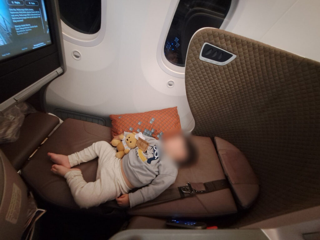 シンガポール航空ビジネスクラスのフルフラットシートで、仰向けにぐっすり眠る1歳くらいの男の子。男の子はお腹にクマのぬいぐるみを抱え、グレーのスウェットと白いパンツを着用している。広い座席スペースと、窓から見える夜景、機内エンターテインメントのスクリーンが映っている。
