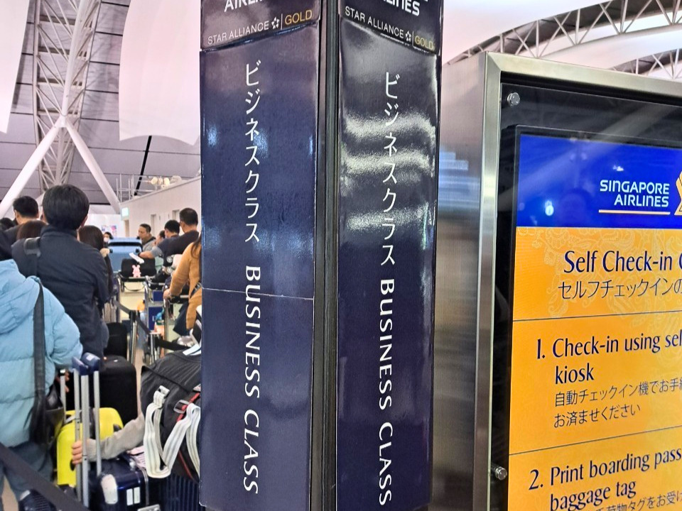 シンガポール航空のビジネスクラス・スターアライアンスゴールド優先チェックインカウンターに並ぶ列。手前には紺色の優先搭乗案内ポールが立ち、奥にはスーツケースを持った旅行客と、オレンジ色の「Self Check-in」案内スクリーンが見える。空港での旅の始まりの様子。
