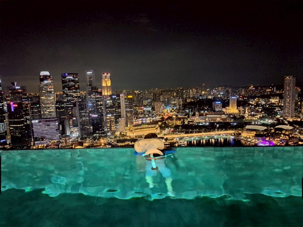深夜のインフィニティプールにて。シティ側の煌びやかな高層ビル群の夜景を背景に、白いライフジャケットを着用した男の子がプールに入り、非現実的な絶景を静かに眺めている背中。