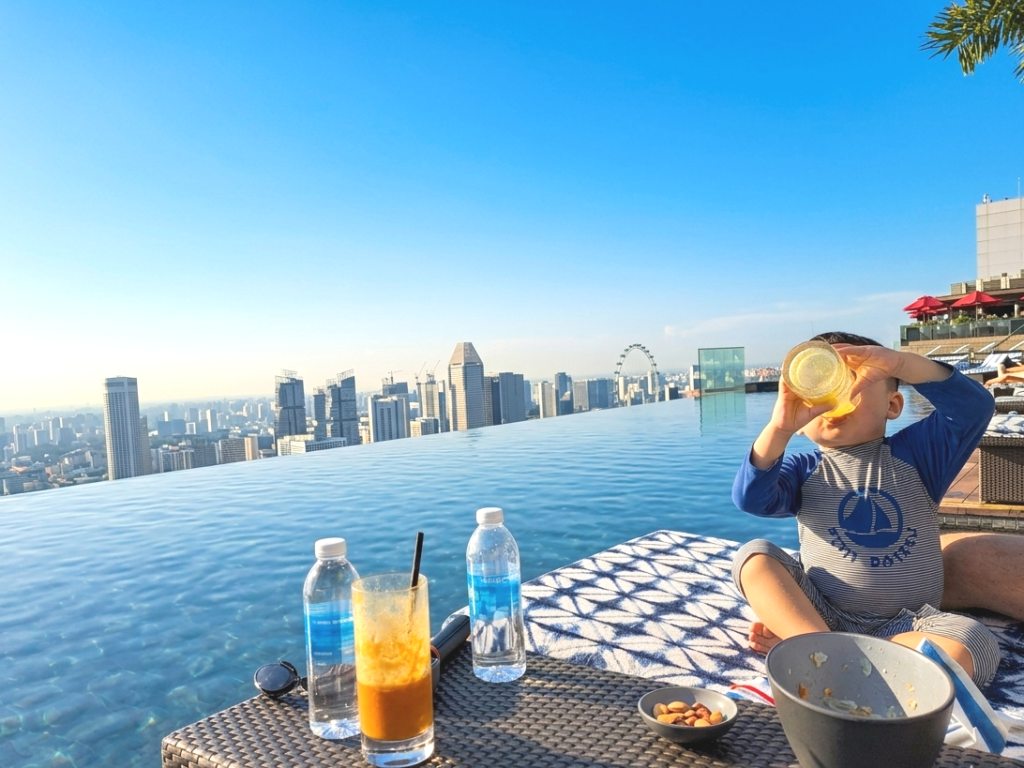 日差しが心地いいプールサイドのウッドデッキにて。水着姿の男の子がデッキチェアに座り、スムージーを飲んでいる。テーブルの上にはカクテル、お水、ポテトが並び、遠くにはシンガポールの景色が見える。