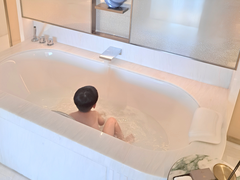 白い大理石が美しいラグジュアリーなバスルーム。ゆったりとした大きな楕円形の白いバスタブに温かいお湯が注がれており、3歳の男の子が気持ちよさそうにお風呂に入っている後ろ姿。