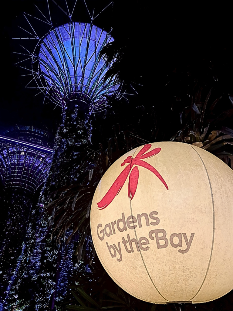 夜のガーデンズ・バイ・ザ・ベイ。手前に「Gardens by the Bay」のロゴとトンボのイラストが描かれた、温かみのあるオレンジ色に光る大きな球体オブジェクトがあり、背景には青や紫の光で鮮やかにライトアップされた巨大なスーパーツリーがそびえ立っている。